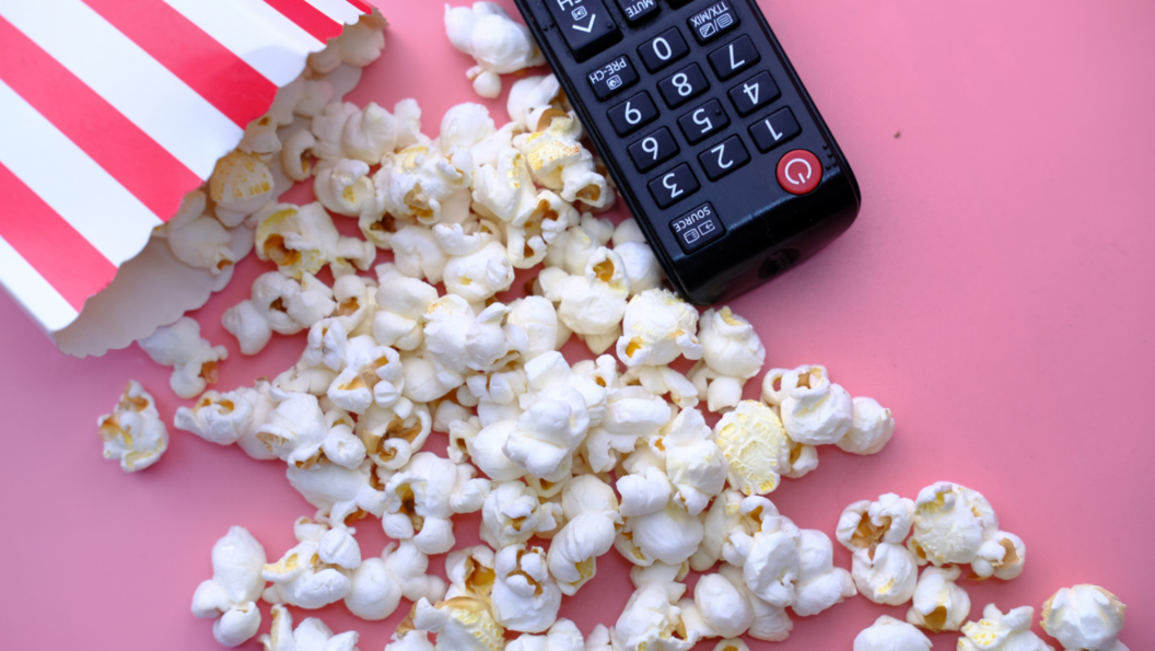 Popcorn in rot-weiß gestreifter Tüte auf rosa Untergrund, daneben schwarze Fernbedienung
