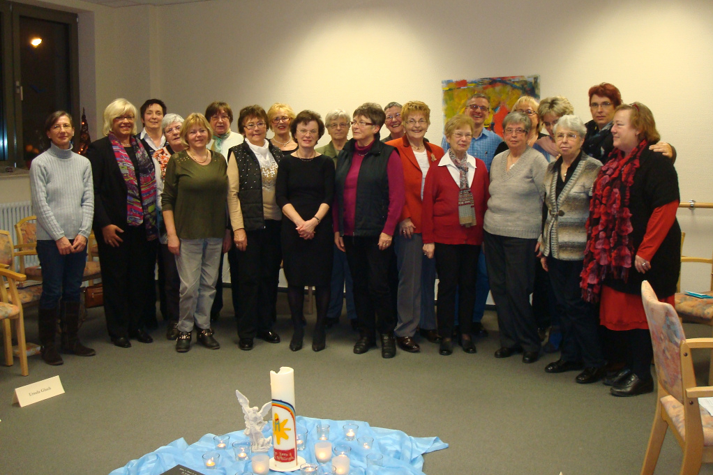 Gruppe von Personen steht in einem Raum vor einer Wand mit einem bunten Bild, im Vordergrund ein Tisch mit blauer Decke und Kerze.