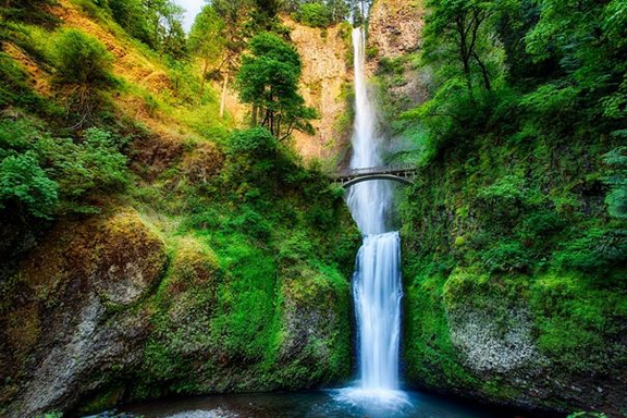 Wasserfall mit zwei Kaskaden und einer Brücke dazwischen, umgeben von grünen Bäumen und Felsen.